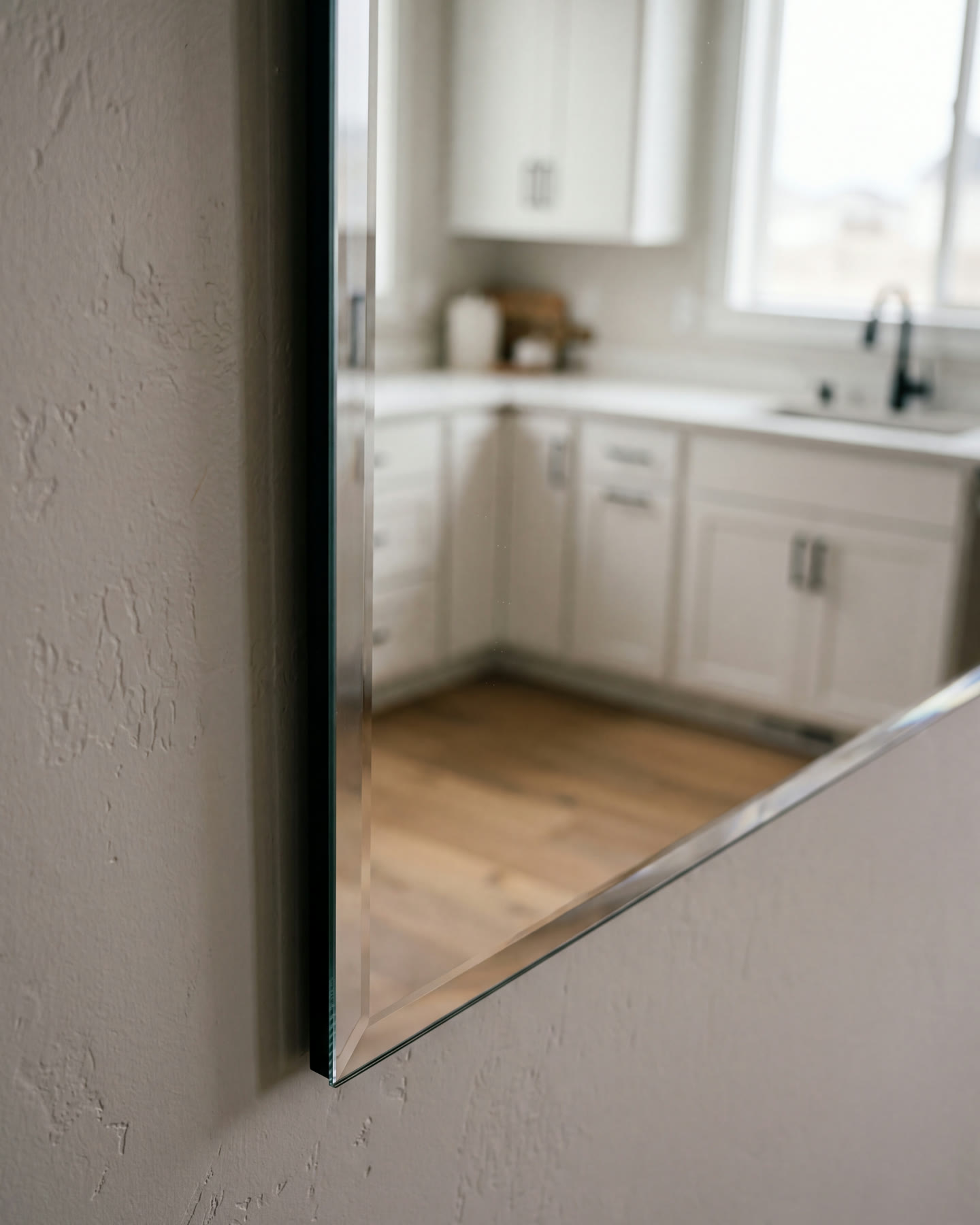 Polished bevelled edge of a frameless wall mirror reflecting a bright kitchen
